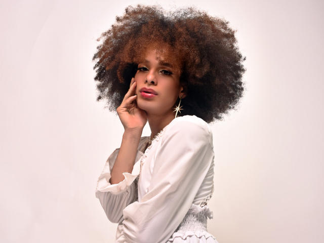 KataleyaFowler with big, fluffy afro, wearing a white blouse, touching her cheek with one hand, star earring, soft smile, light skin, and pink lipstick.