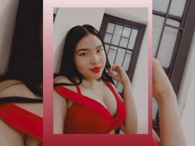 OliviGrace with red bra, showing her tits, smiling with red lipstick, black hair, holding bra strap, in a photo with a pink border, window in background.