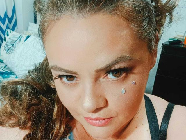 Vanessa Deluca, com cabelo castanho encaracolado, olhos castanhos, delineador preto, pequena piercing facial em formato de diamante abaixo do olho direito, alça preta, batom rosa suave e um leve sorriso..