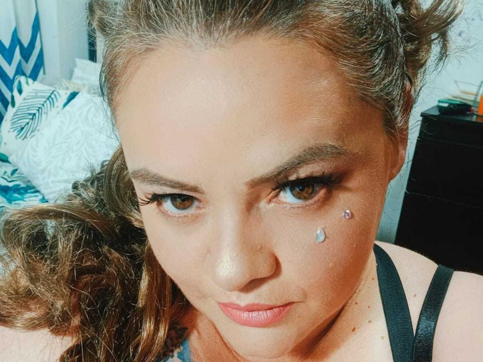 Vanessa Deluca, met krullend bruin haar, bruine ogen, zwarte eyeliner, een kleine diamantpiercing onder het rechteroog, een zwarte bandje, subtiele roze lippenstift en een lichte glimlach..