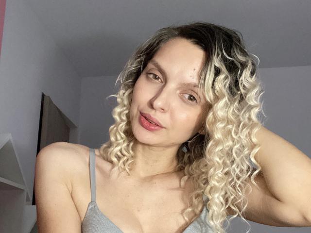 DouceAmelia, con cabello rubio rizado, camiseta gris y labios rosados, mirando a la cámara con una ligera sonrisa, con la mano en el pelo, mostrando pequeños senos y un rostro lindo..