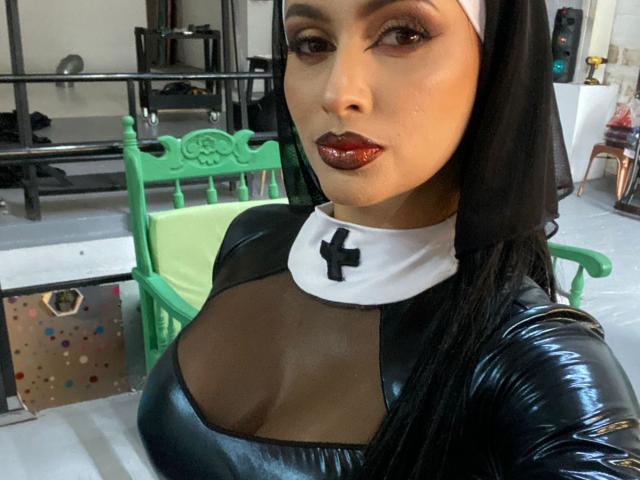 EmilyParkerr, con una top in lattice nero lucido, grandi seni, un copricapo da suora, rossetto rosso scuro e lunghi capelli neri, in piedi in una stanza con una sedia verde e scaffali in metallo..
