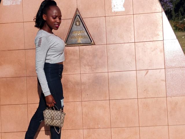 LoverGia, avec sa peau foncée, son rouge à lèvres rouge et ses cheveux tressés, porte un crop top gris, un jean noir et un sac à main matelassé, et se tient près d'une plaque triangulaire sur un mur recouvert de carreaux pêche..