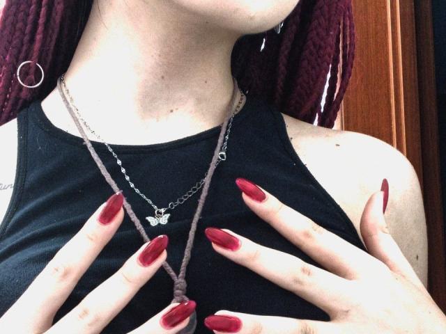 Les mains de Zanoza, avec ses ongles rouges, couvrent ses seins, elle porte un haut noir, un collier en chaîne et des boucles d'oreilles cercles, avec des dreadlocks marron, un teint clair et un collier avec un papillon..
