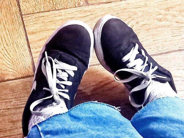 Les baskets noires de Zanoza, avec des lacets blancs et des bords effilochés, posées sur un plancher en bois, avec un jean bleu légèrement déchiré..
