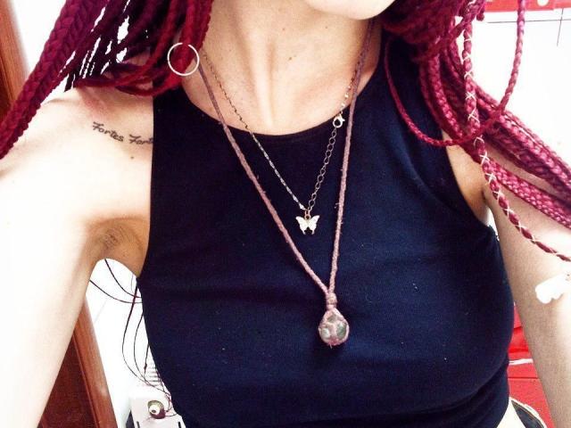 Zanoza tem tranças vermelhas, uma blusa preta sem mangas e usa um colar com uma borboleta e um pingente de pedra transparente. Seus seios estão visíveis e ela tem uma tatuagem..