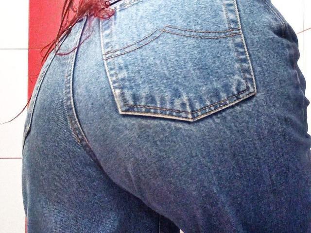 Zanoza's blue denim ass, showing a prominent back pocket and tight, worn-in jeans. Red hair partially visible. Buttocks are large and slightly curved.