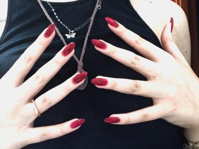 Las manos de Zanoza, con uñas rojas y brillantes, tocan su camiseta negra, cubriendo sus pechos. Lleva un collar con forma de mariposa y un anillo sencillo en su mano derecha..