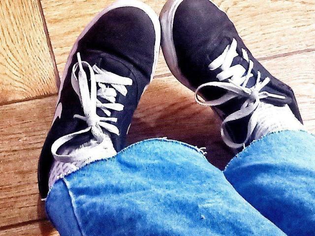Le sneakers nere di Zanoza, con lacci bianchi, leggermente usurate, sopra jeans blu sfilacciati, in piedi su un pavimento di legno. Le sneakers hanno un aspetto casual, leggermente sporco..