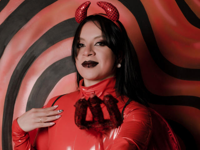 KatalinaJohnson with black hair, red devil horns, glossy red latex outfit, black lipstick, smiling, hand on chest, red and black background, wearing pearl earrings.