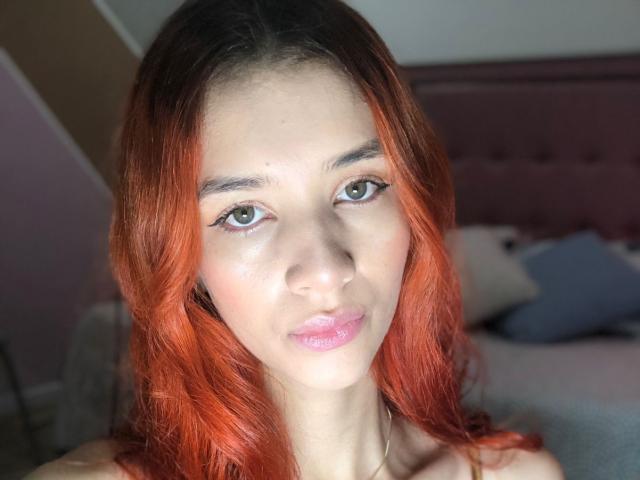Greta Harris heeft fel oranje-rood haar, groene ogen, glanzende roze lippen en een lichte huidskleur. Ze draagt een zilveren ketting en kijkt recht in de camera..
