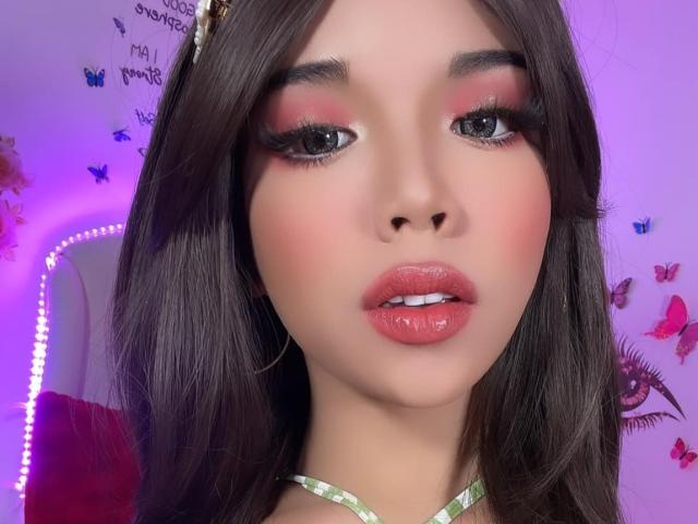 AyeshaSebastian's face, showing glossy pink lips, shiny eyes with heavy makeup, and dark brown hair, with a green choker around her neck. Her ass is not visible.
