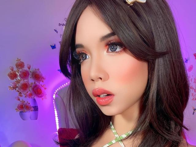 AyeshaSebastian, con lunghi capelli scuri, un leggero rossore sulle guance e labbra rosa lucide, indossa una collana verde a quadretti, ha gli occhi azzurri e un viso carino, con la bocca leggermente aperta..