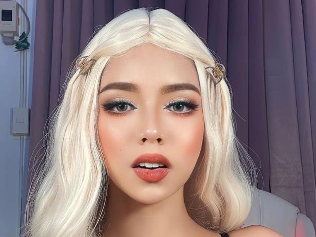 AyeshaSebastian a les cheveux blond platine, les yeux verts et les lèvres pleines roses. Elle porte de petites pinces à cheveux sur les côtés, avec du fard à paupières bleu et un eye-liner épais. Ses seins sont visibles..