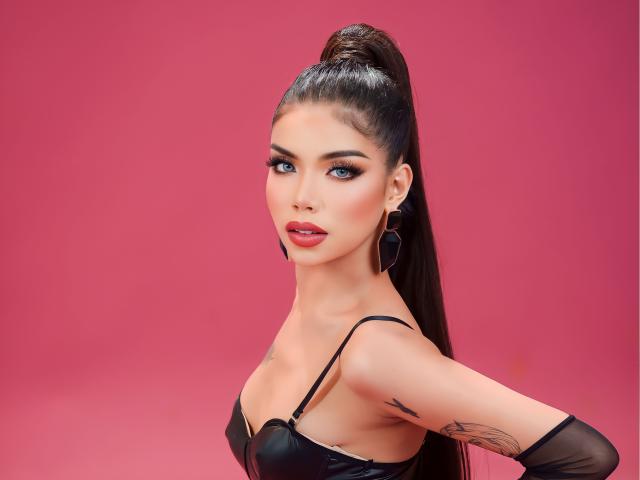 AyeshaSebastian mit hohem Pferdeschwanz, schwarzem Lederoberteil, rotem Lippenstift, Reifenohrringen und Tattoo am Arm, die über die Schulter schaut und die Hände in die Hüften gestemmt hat, vor einem rosa Hintergrund..
