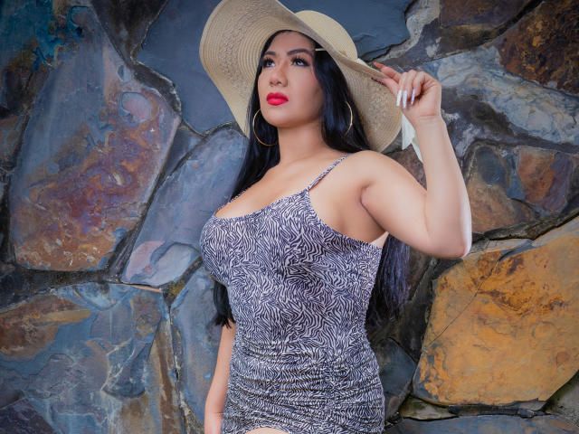 KhloeLawson with big tits in a tight, zebra-patterned dress, holding a beige hat, wearing red lipstick, and large gold hoop earrings, standing against a rocky wall.