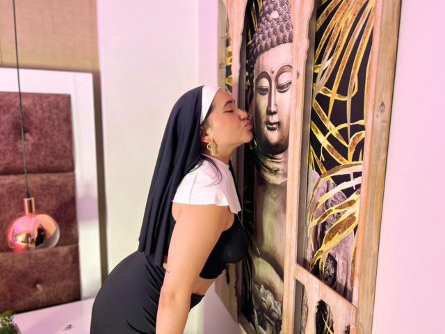 VeronicaWins, vestida con un hábito de monja, besando un cuadro de Buda pintado en un panel de madera. Sus pechos son visibles debajo del atuendo blanco y negro, con los ojos cerrados..