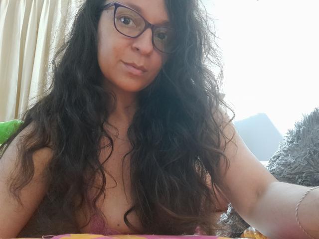 Angelonix with long, wavy black hair, glasses, and a pink lace bra, showing her tits, sitting on a bed, looking at the camera with a slight smile.