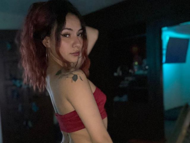 Valery Harllow, avec ses cheveux rosâtres, porte un haut rouge sans bretelles et on peut voir un tatouage sur son épaule droite. Elle regarde par-dessus son épaule avec un léger sourire. Ses seins sont visibles..