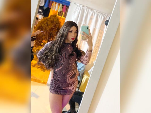 CherilynDoll with long brown hair, wearing a tight, swirling purple dress, holding a phone in a mirror selfie, showing her ass and a slight butt cheek.