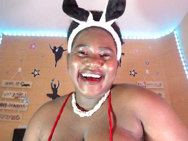 Dilay with dark skin, smiling, wearing white bunny ears, red halter top, and white bead necklace, showing shiny face and red lips, in a brightly lit room.