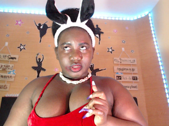 Dilay with dark skin wearing a red dress, white headband with black bunny ears, and white beaded necklace, holding a white and purple striped dildo, looking at camera.