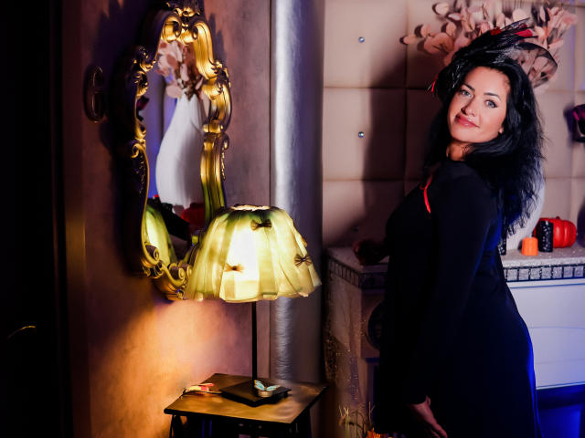 Anelisa Reys, con cabello negro y rizado, vestida con un vestido negro, de pie junto a un espejo dorado, una lámpara amarilla y una mesa de madera, mostrando su trasero, sonriendo, en una habitación con poca luz..