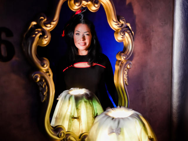 AnelisaReys, con i capelli neri e un dettaglio rosso su una maglia nera, sorride, di fronte a uno specchio dorato decorato, con due paralumi verdi che emettono una luce soffusa..