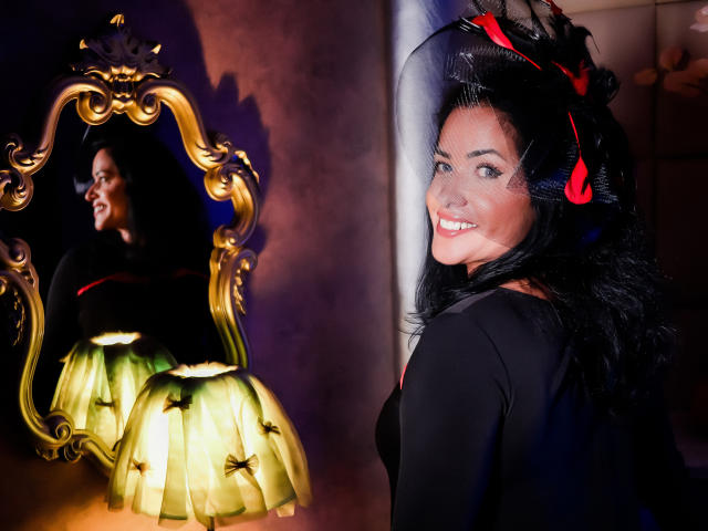 AnelisaReys, avec ses cheveux noirs, son chapeau rouge et sa robe noire, sourit en se regardant dans un miroir qui la reflète. Elle a des yeux pétillants et un nez mignon, et il y a une lampe en arrière-plan..