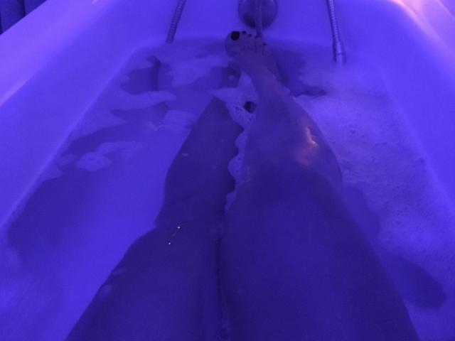 Les jambes d'EstrelaFrenchie dans une baignoire éclairée en violet, avec les pieds partiellement immergés dans de l'eau savonneuse, les orteils visibles, avec du vernis à ongles noir, de l'eau et des bulles autour..