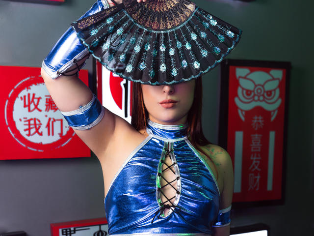 LaiaReyd porte une robe en latex bleu brillant avec un laçage sur le devant, tenant un éventail métallique devant son visage, et portant des manchettes assorties. Ses seins sont à peine visibles sous l'éventail..