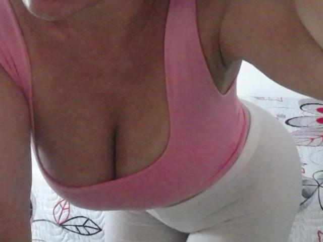 VictoriaKim's tits in a pink tank top, showing ample cleavage. She's wearing white pants, leaning forward, and the photo is taken from a high angle.