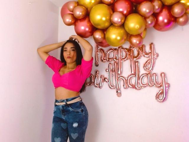 SarahyJhones, con i capelli castani, un top corto rosa e jeans blu con motivo a cuori, è in piedi con le mani tra i capelli, accanto a palloncini dorati e rosa, mostrando i suoi seni e i suoi glutei..