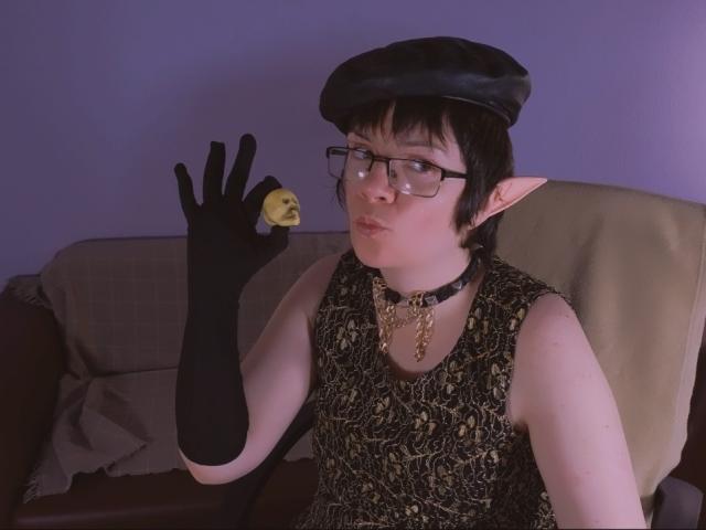 EverlyRays with elf ears, black gloves, flat cap, glasses, and chain collar, holding a yellow lemon, pouting, wearing a snake-pattern dress, seated on a beige chair.