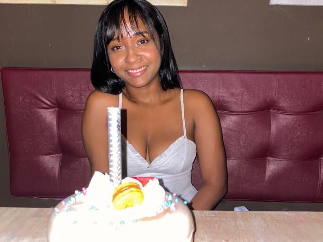 Melany Scoth, mit dunklem Haar und mittelbrauner Haut, lächelt in die Kamera und trägt eine weiße Spitzenbluse, die ihre Brüste zeigt. Sie sitzt auf einem Stuhl, vor ihr steht eine Torte..