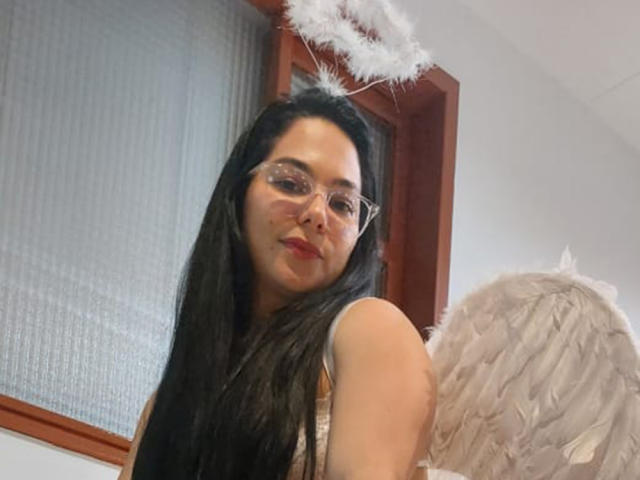 Bianca Scott, com cabelos pretos longos, óculos e um halo, vestindo asas de anjo brancas, com um sorriso discreto, aparecendo de costas em uma selfie..