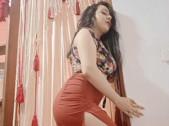 MabyLegrand with long black hair, floral top, and tight red skirt, showing her ass and side boob, leaning against a wall with a confident, slightly seductive look.