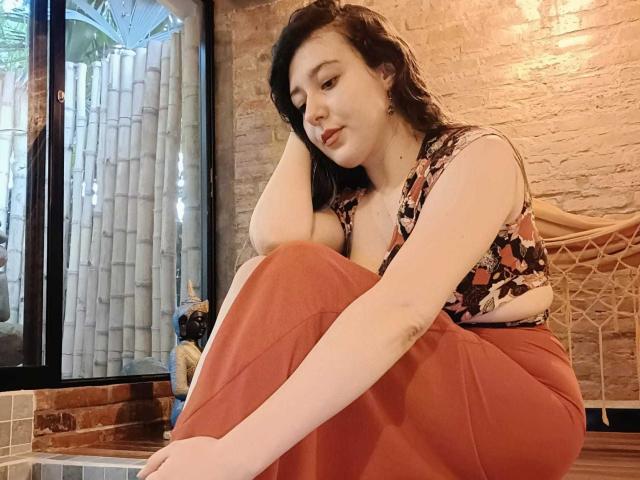 MabyLegrand with pale skin, dark wavy hair, wearing a floral top and red pants, sits with arms and legs crossed, head resting on hand, looking down. Soft, cute.