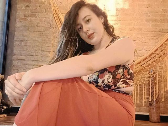 MabyLegrand with long brown hair, wearing a floral tank top and red skirt, sitting with arms and knees together, looking at the camera, in a cozy room.