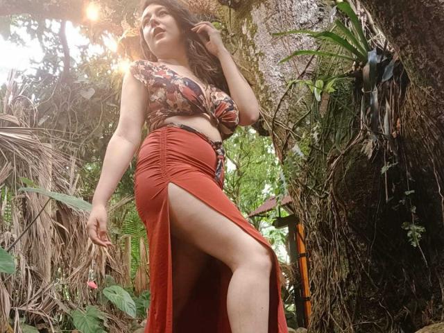 Maby Legrand, con grandes senos, vistiendo una camiseta corta con estampado floral y una falda roja con una abertura alta en la pierna, de pie en un entorno que se asemeja a una jungla, mirando hacia arriba con una ligera sonrisa..