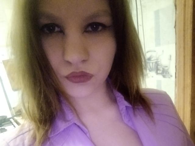 Lisanna, avec ses cheveux bruns clairs, son t-shirt violet et son rouge à lèvres rose, regarde directement l'appareil photo avec une expression sérieuse, laissant légèrement entrevoir ses seins à travers le t-shirt..
