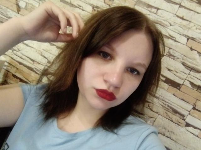 Lisanna, com cabelo castanho, batom vermelho e camisa azul clara, segurando uma pequena pedra perto da cabeça. Seus seios são pequenos e suas nádegas não estão visíveis..