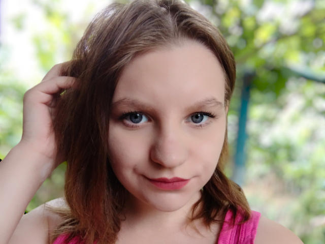 LisannaHot heeft blauwe ogen, een lichte huid en bruin haar, en draagt een roze top. Ze glimlacht en heeft haar hand in haar haar. Haar borsten zijn klein, en ze is schattig..