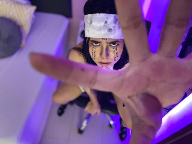 LianaBernocchi with black hair, white headband, face paint, looking up, hand in foreground, sitting on chair, wearing black lingerie, purple light, braided hair, serious expression.