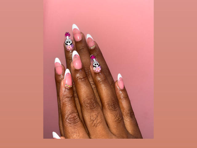 MelanyScoth's hands with dark brown skin, long pink and white French tip nails, small pink flowers, and skull designs on the nails. She's showing off her manicured fingers.