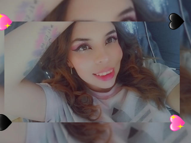 VioletaStone tem cabelos castanhos ondulados, sombra rosa nos olhos e está sorrindo com os lábios rosados. Ela está vestindo uma camisa cinza clara, e parece adorável, com os seios levemente visíveis..
