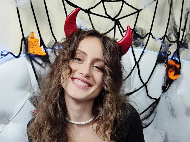 Alesya Drons, con i capelli castano ricci, corna da diavolo rosse, che sorride, indossa una maglietta nera e una collana di perle bianche, e guarda leggermente di lato..