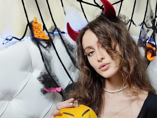 Alesya, com cabelos castanhos ondulados, usando chifres vermelhos de diabo, segurando uma máscara amarela, uma blusa preta e um colar de prata, sentada em um sofá branco com estofamento..