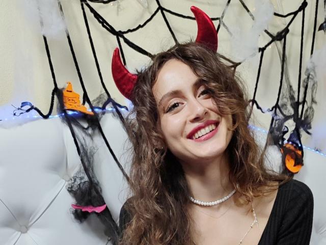 Alesya Drons mit lockigem, braunem Haar, roten Teufelshörnern, pinkem Lippenstift, lächelnd, trägt ein schwarzes Oberteil und eine Perlenkette und zeigt ihre weißen Zähne. Orange und schwarze Halloween-Dekorationen..