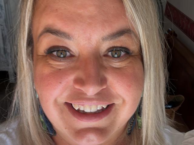El rostro de BlueMaryAnne, mostrando cabello rubio claro, ojos azul verdosos, maquillaje sutil y una sonrisa con dientes visibles, lleva aretes coloridos, en un primer plano con iluminación suave..